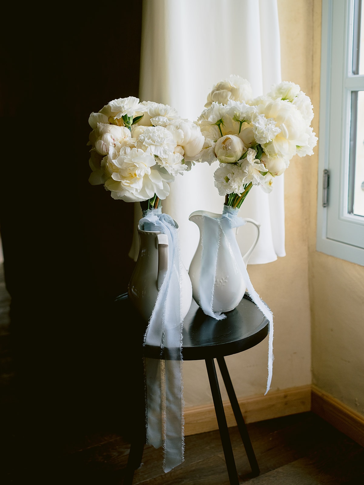 Bouquets de pivoines et oeillets blancs dans des brocs en ceramique blanche noues d un ruban bleu ciel devant un rideau blanc - Bouquetier Paris 17